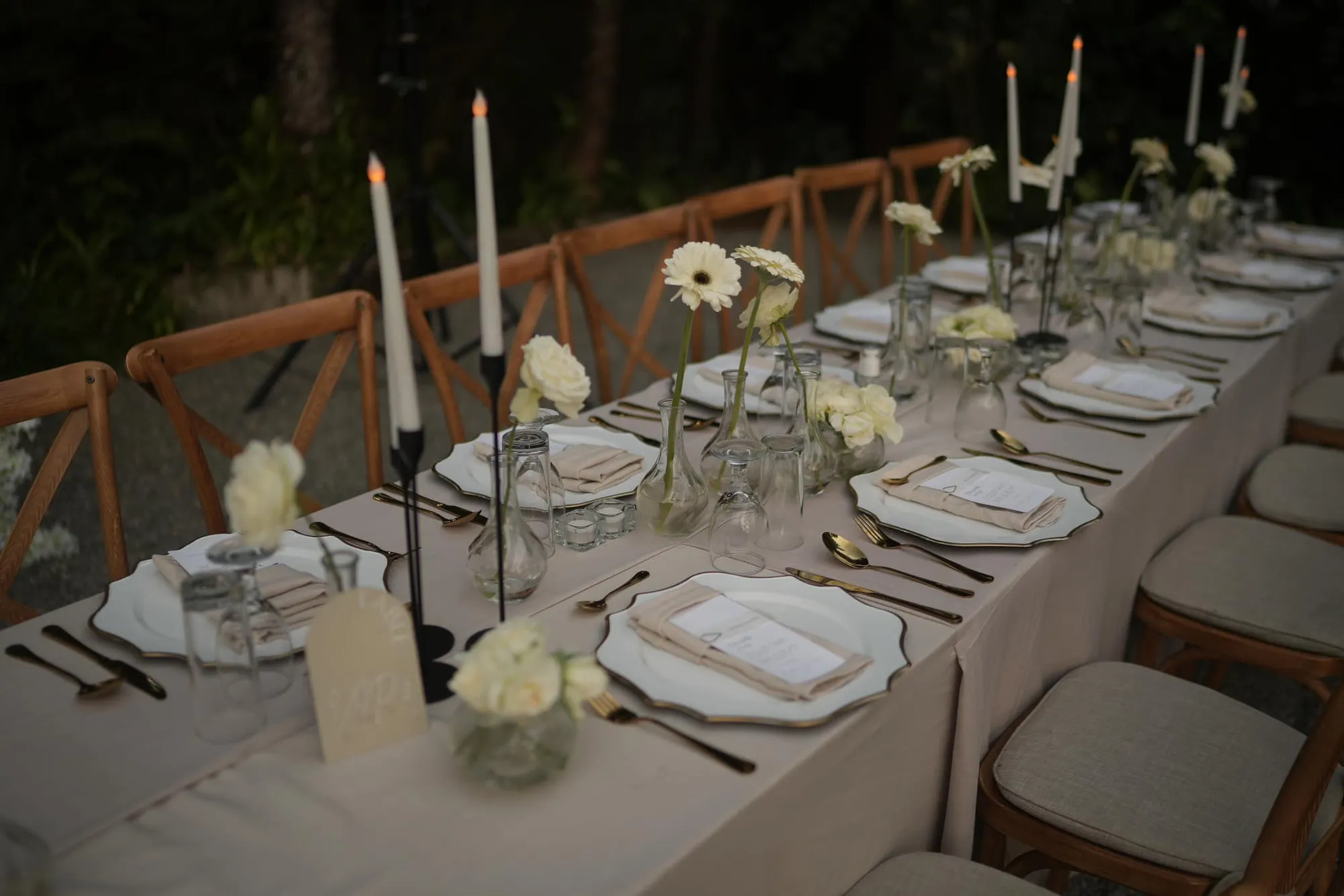 Long reception table with cream linens, gold cutlery, white gerberas in bud vases, and black candlesticks holding white tapers.