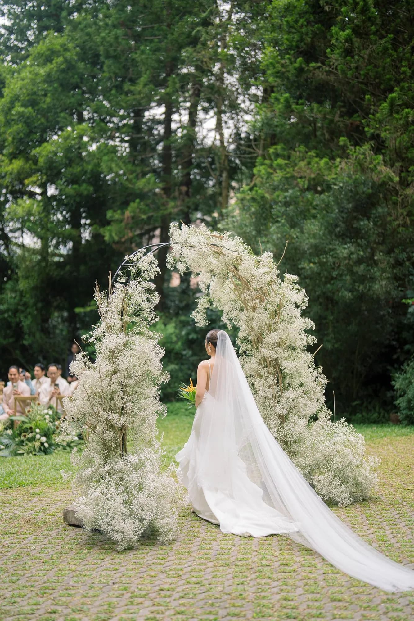 Bride with a long veil walking through a crescent-shaped baby's breath arch on a cobblestone lawn.