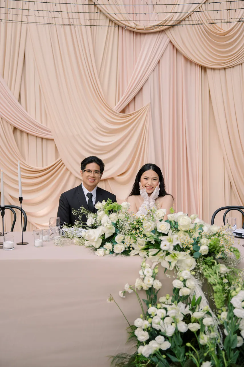 Couple seated at the head table before a swagged peach drape backdrop, with white roses and chamomile cascading along the table edge.