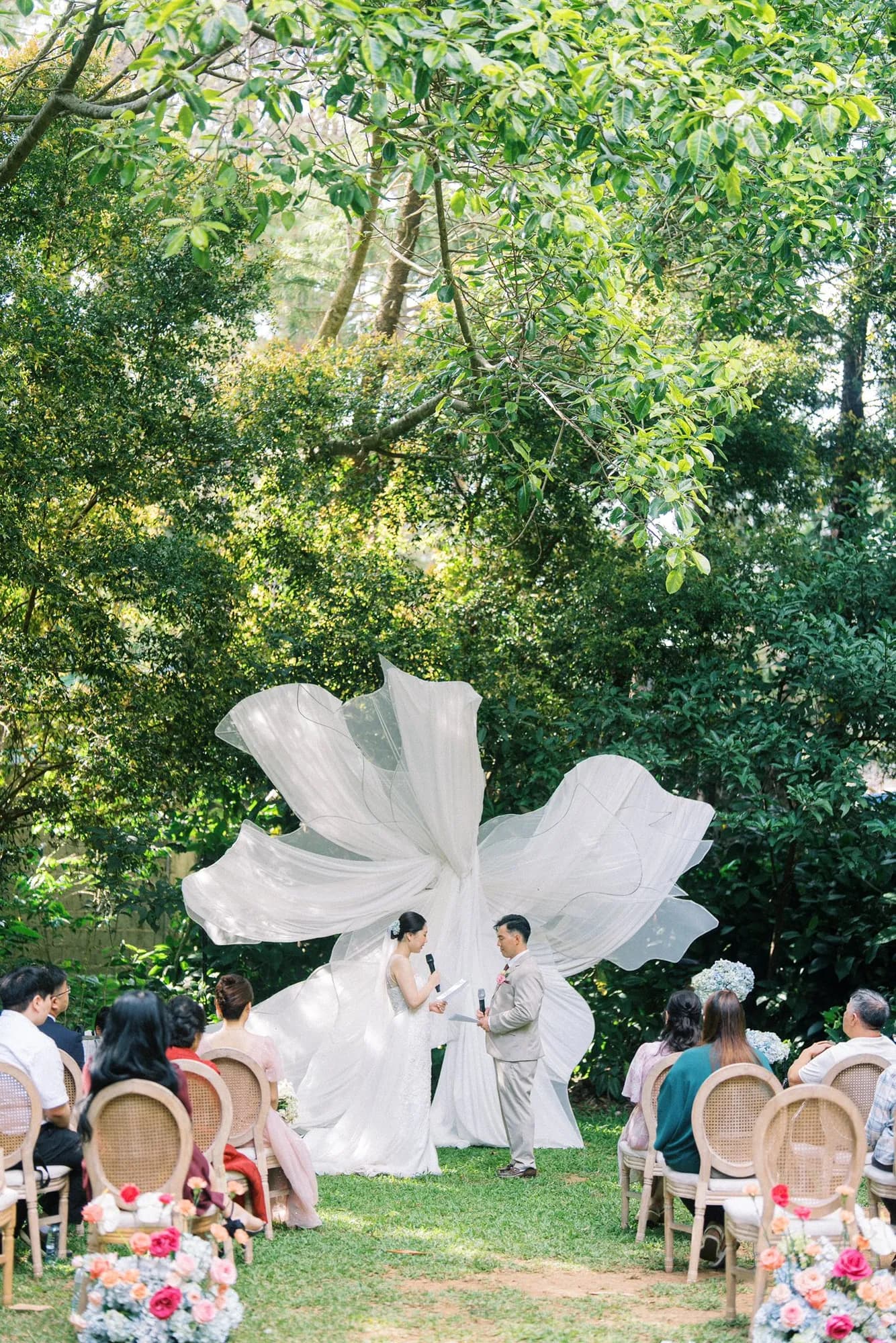 Couple exchanging vows before a sculptural white tulle installation, with guests seated on cane-back chairs.