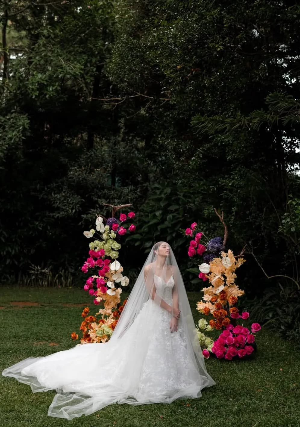 Bride in a long veil standing between asymmetric floral pillars of fuchsia, orange, green, and lilac blooms.