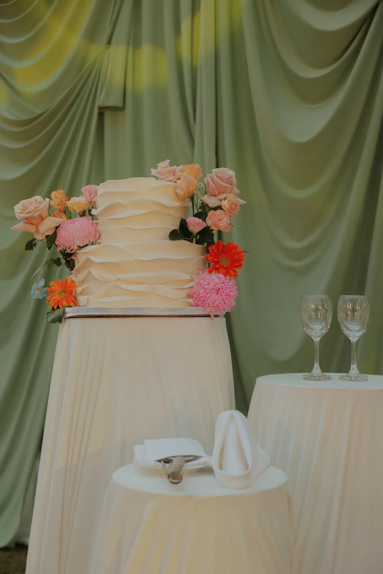 Two-tier ruffled white wedding cake dressed with peach roses, pink chrysanthemums, and orange gerberas against sage-green drapery.