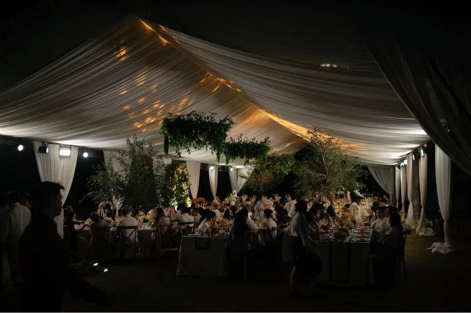 Night view inside a white-draped tent with suspended greenery, warm uplighting, and guests dining at flower-filled tables.