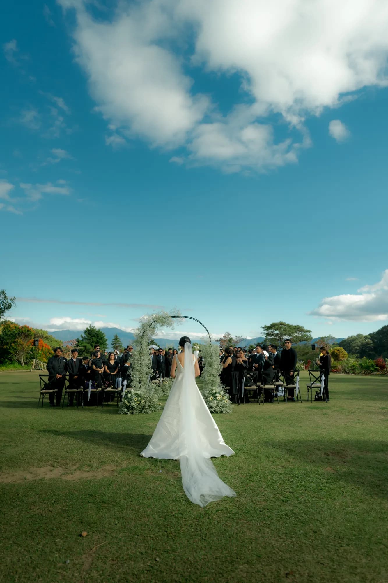 Bride walking down the grass aisle toward a baby's breath arch, with guests in black and mountains on the horizon.