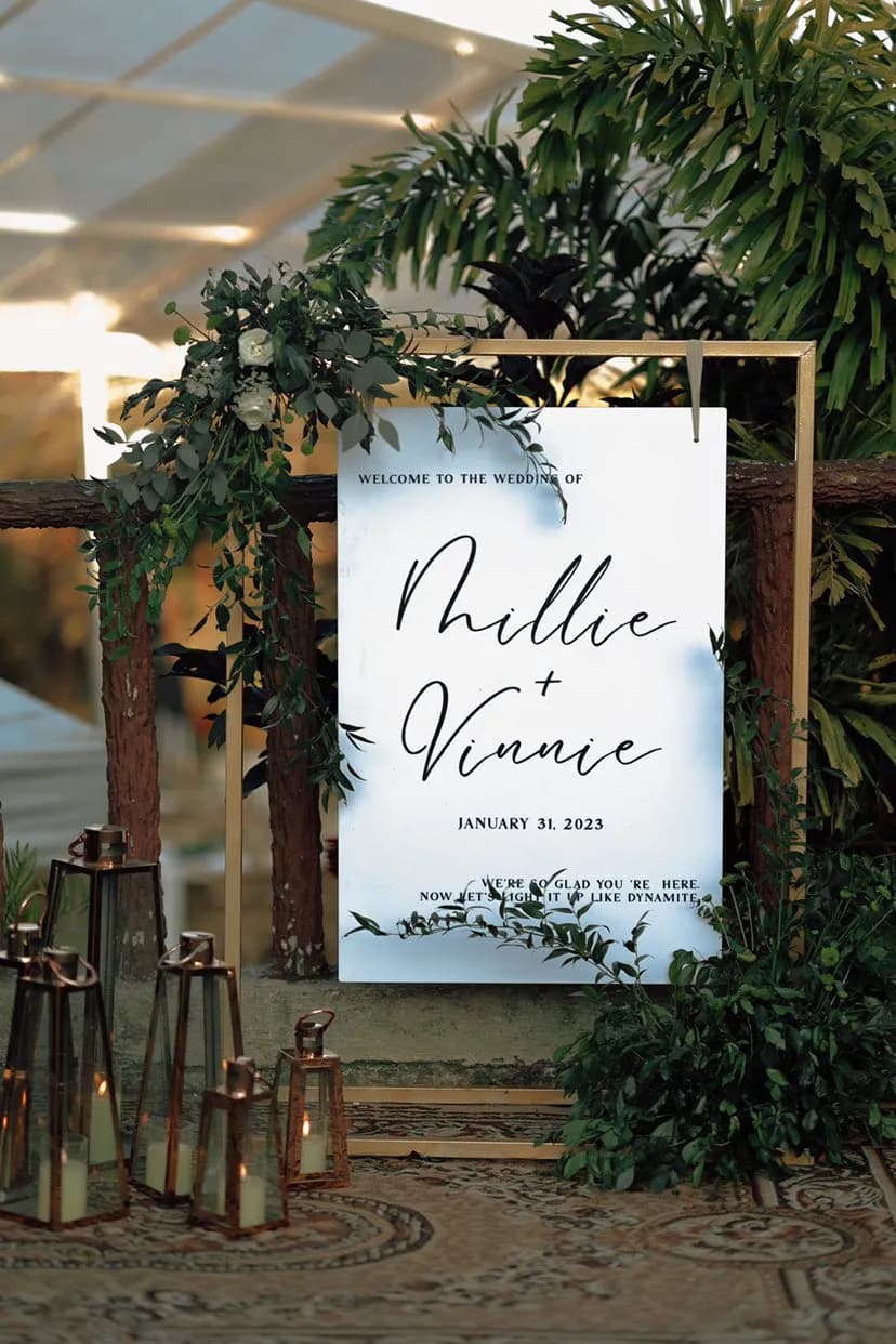 White welcome signage in a gold frame draped with greenery and white blooms, with copper lanterns clustered below.