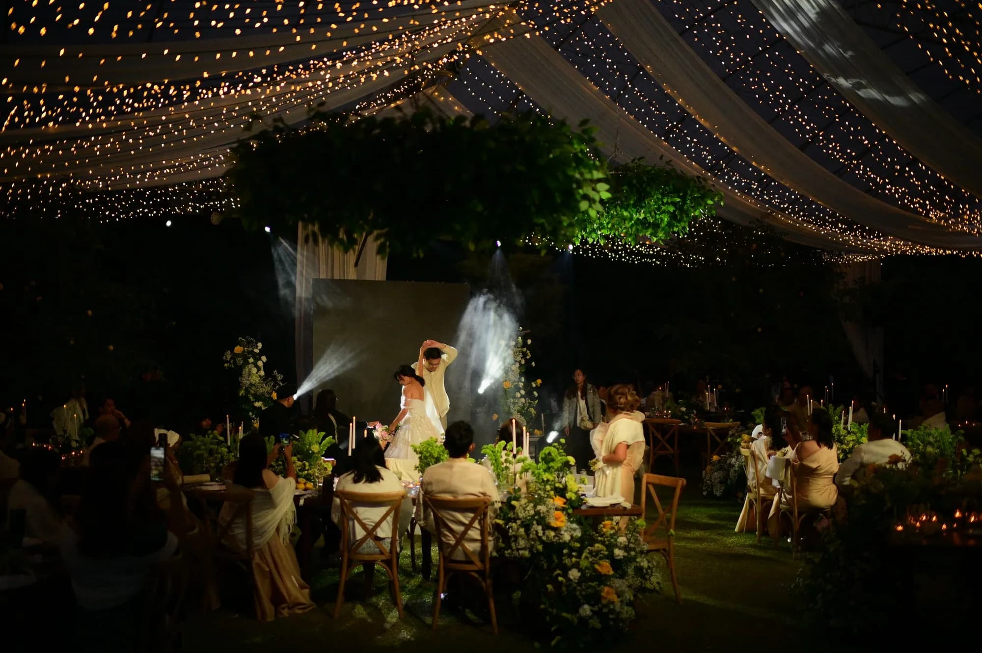 Couple's first dance under a fairy-light canopy and greenery chandeliers, surrounded by guests and green-and-yellow florals.