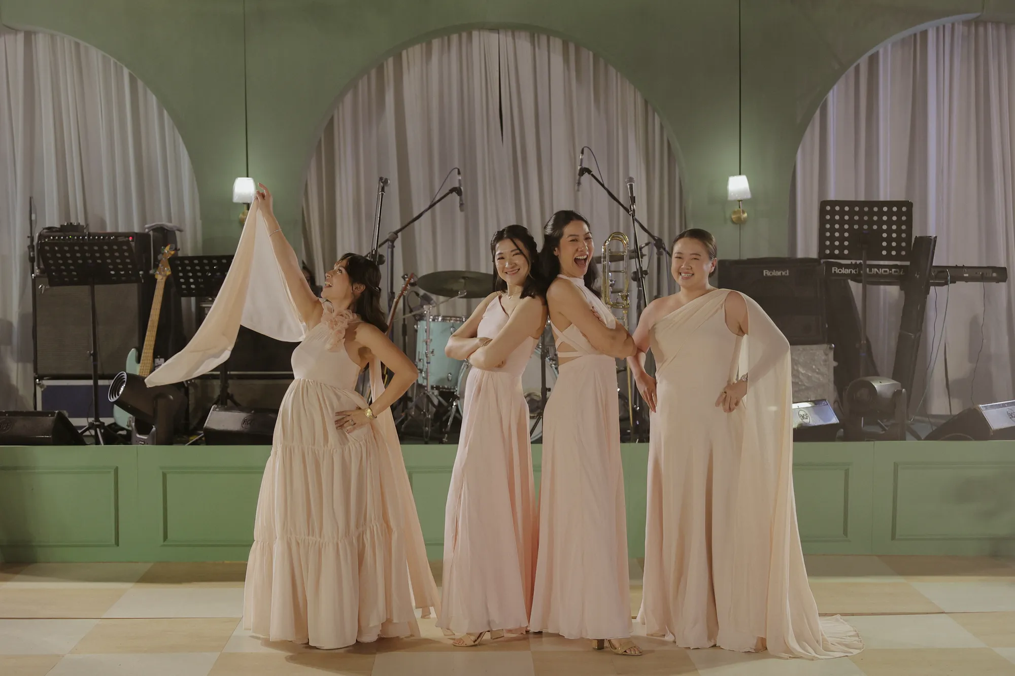 Four women in blush gowns posing on a checkerboard dance floor in front of a sage-green arched stage set for a band.