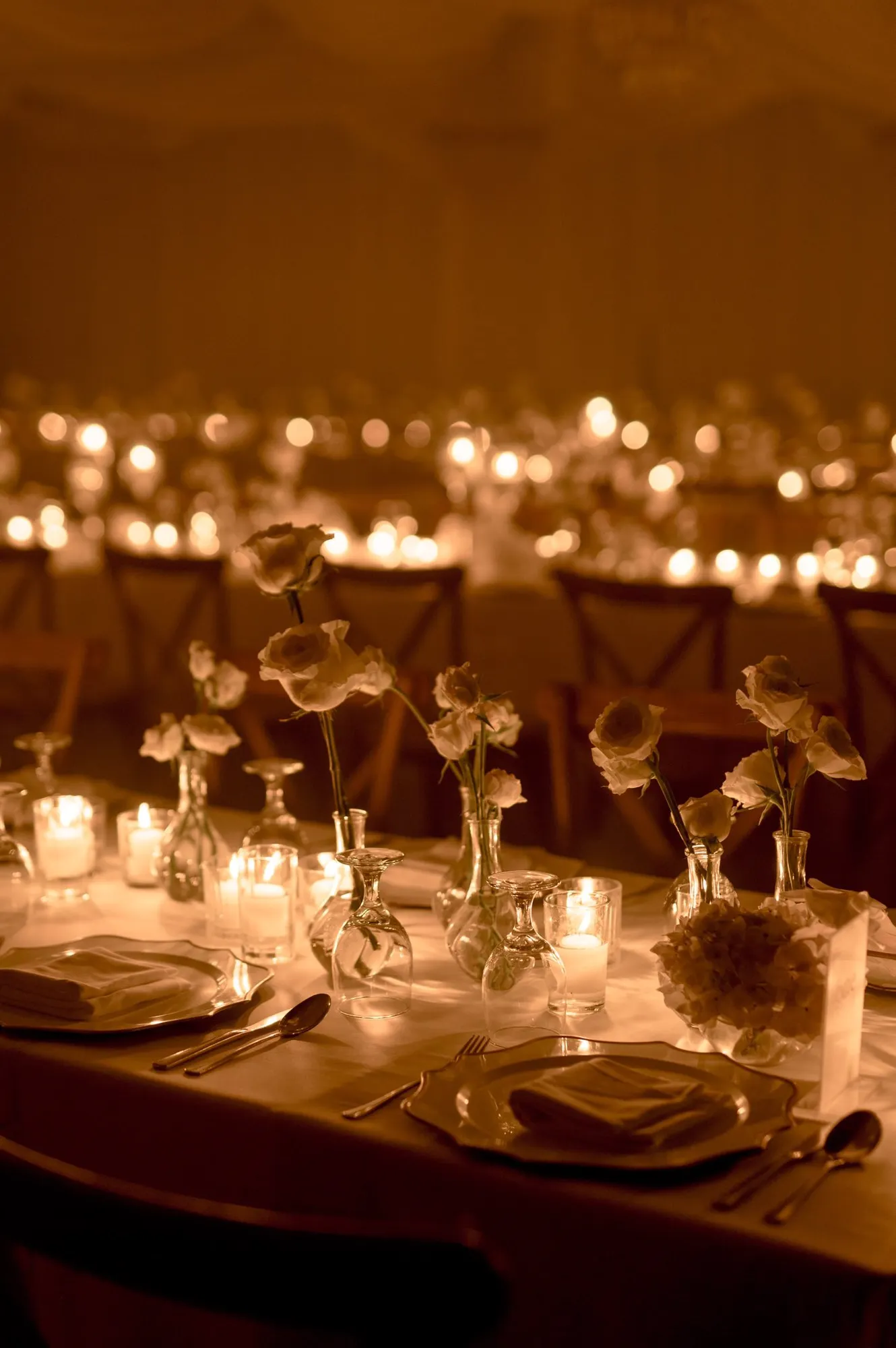 Long candlelit reception table with single-stem white roses in bud vases and gold-rimmed chargers.