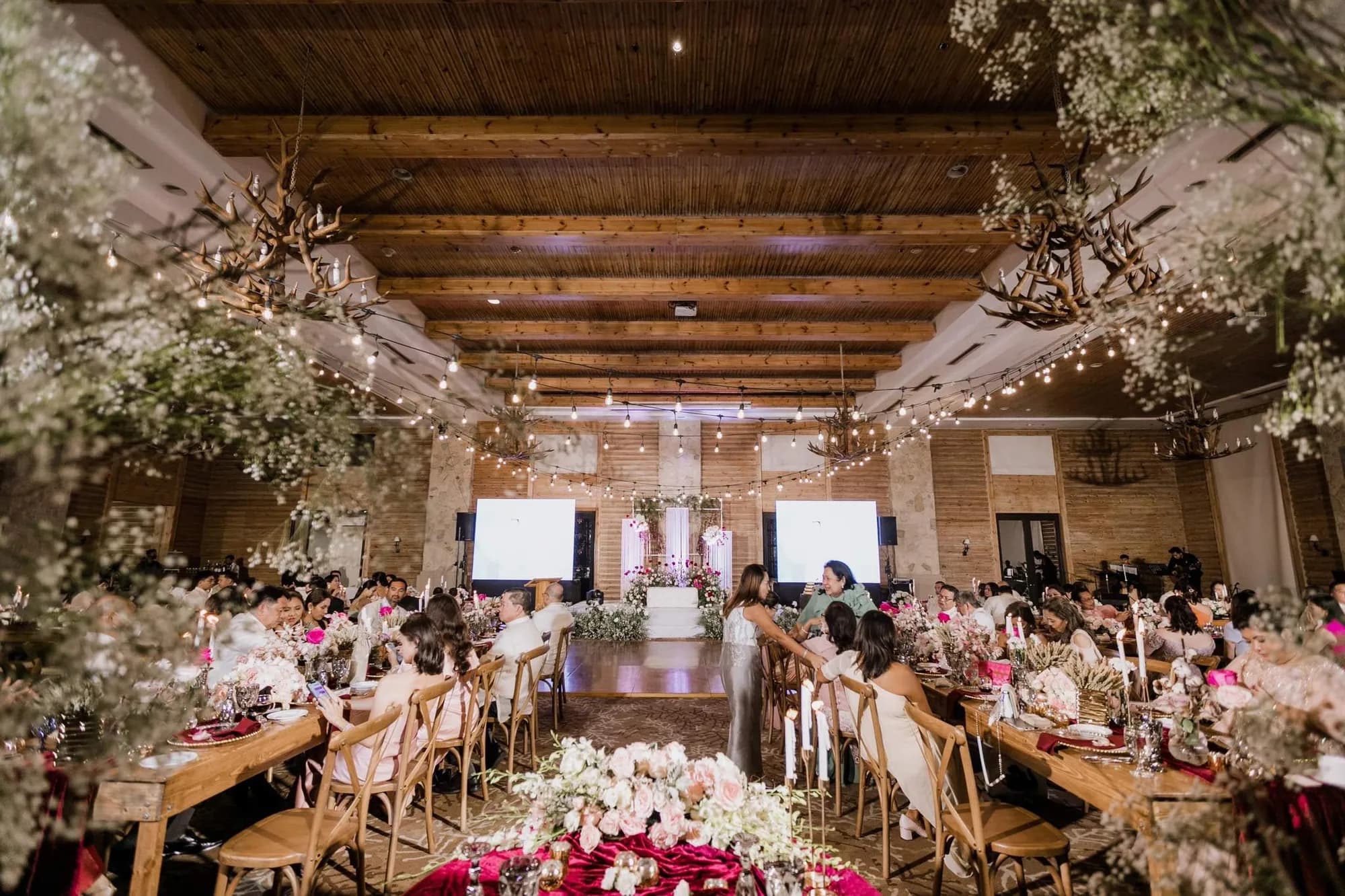 Timber hall with antler chandeliers, string lights, hanging baby's breath, and guests seated at long wooden tables.