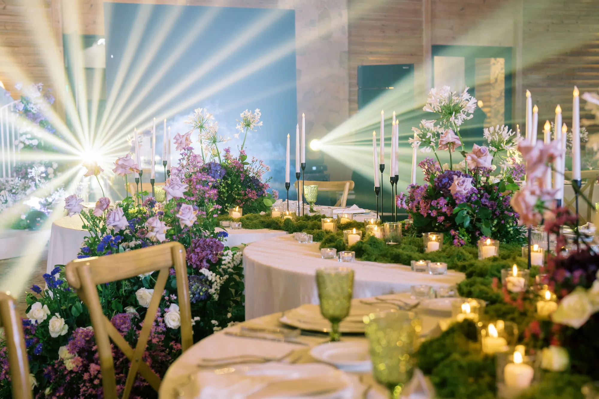 Serpentine tables edged in moss with purple wildflowers, taper candles, and beams of light across the room.