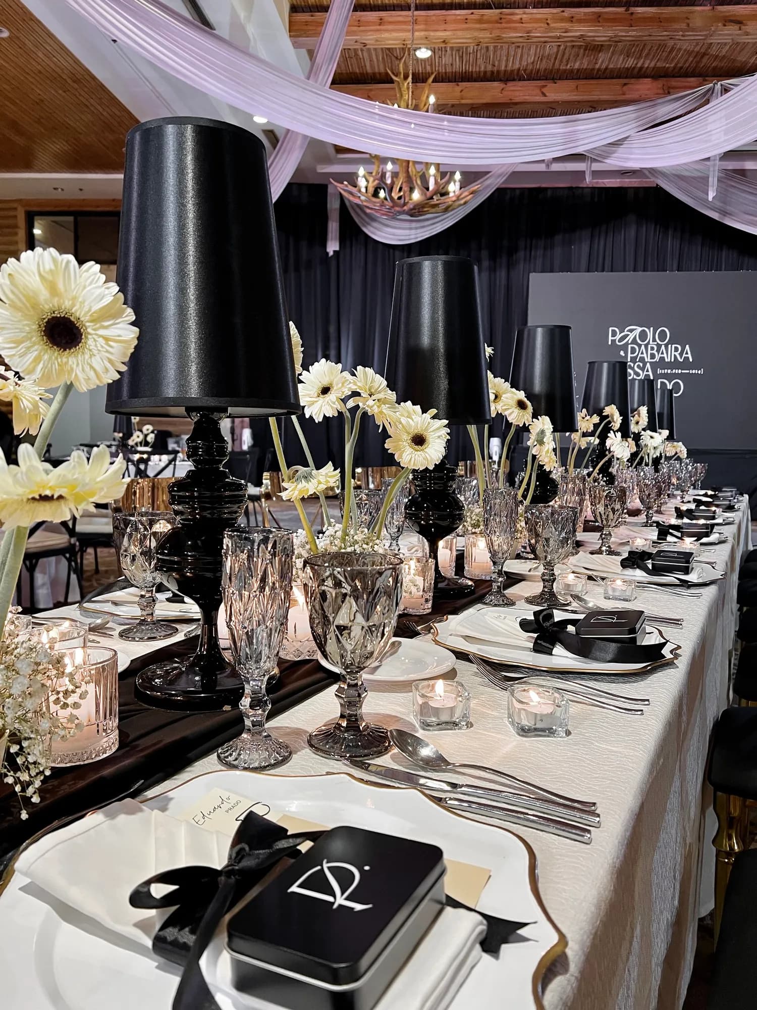 Long table with black lampshade lamps, cream gerbera daisies, smoked glass goblets, and black-ribboned favor tins.