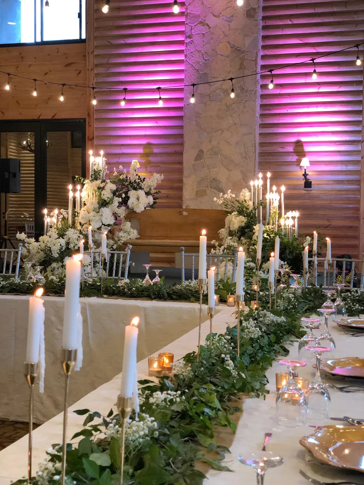 Head table with greenery garland, gold taper candlesticks, and white florals against pink-lit timber walls.