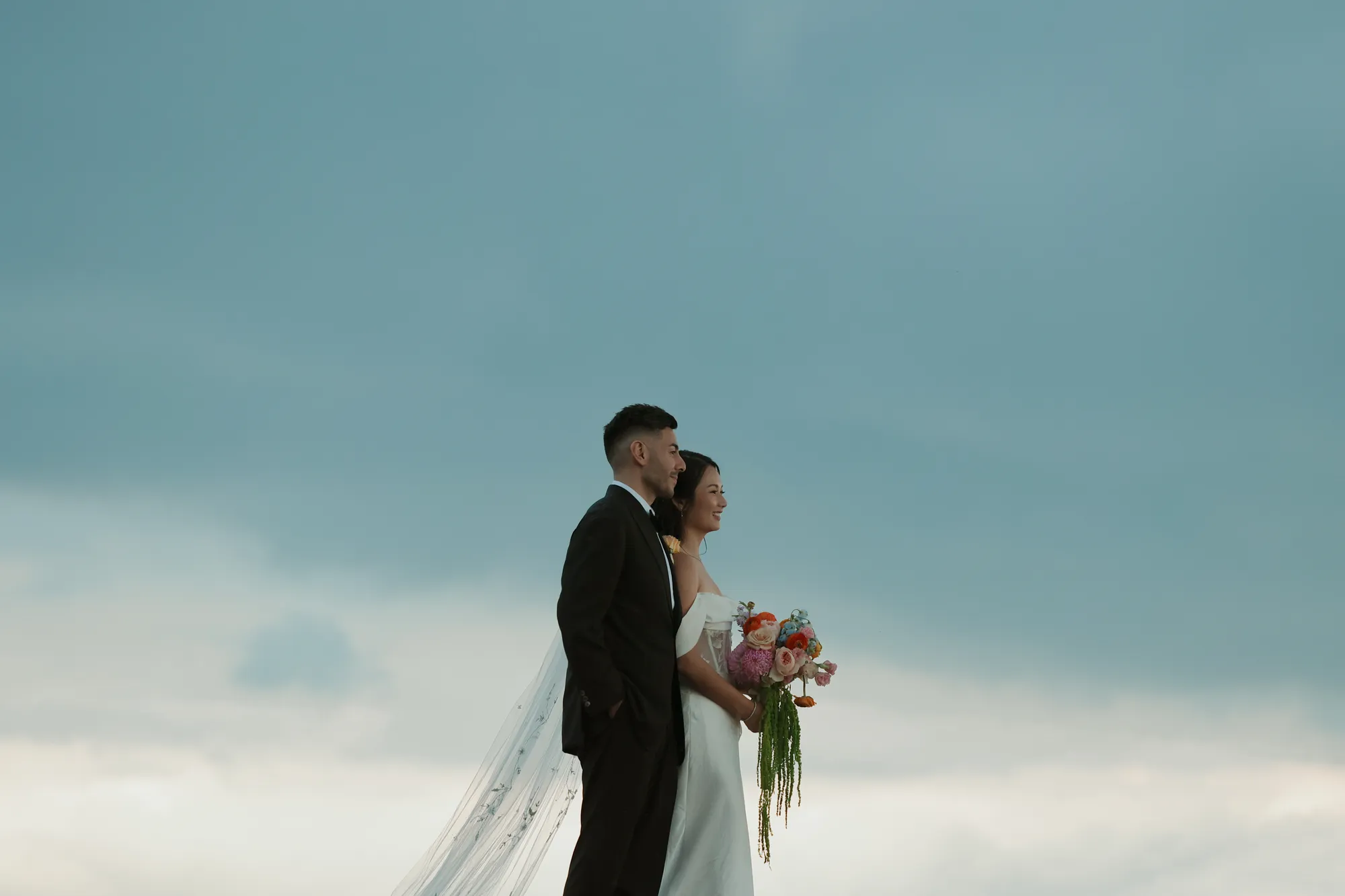 Bride and groom stand in profile against a wide blue sky, the bride holding a bouquet of pink, peach, orange, and blue blooms with trailing amaranthus.