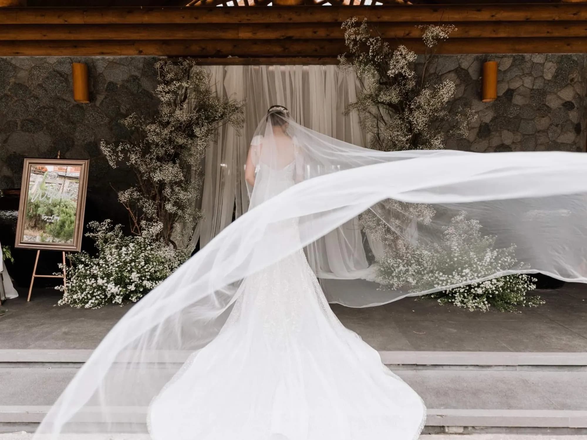Bride's veil sweeps across the frame at the chapel entrance, styled with baby's breath trees, ground clouds of gypsophila, sheer drapery, and a framed mirror welcome sign.