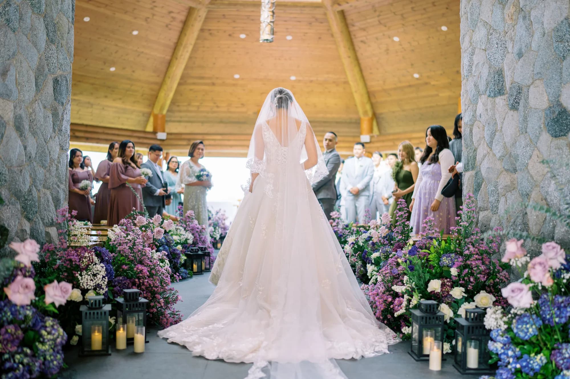Bride in a cathedral veil stands at the head of the aisle between lush beds of lavender, purple, and blue florals with black candle lanterns.