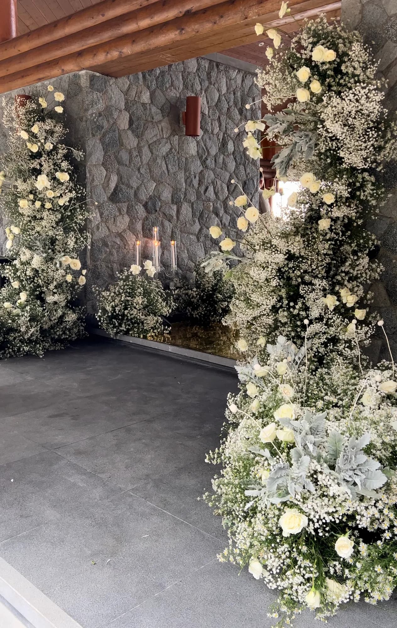 Towering arrangements of baby's breath, cream roses, and dusty miller frame a stone wall where a crystal candelabra with lit taper candles glows.