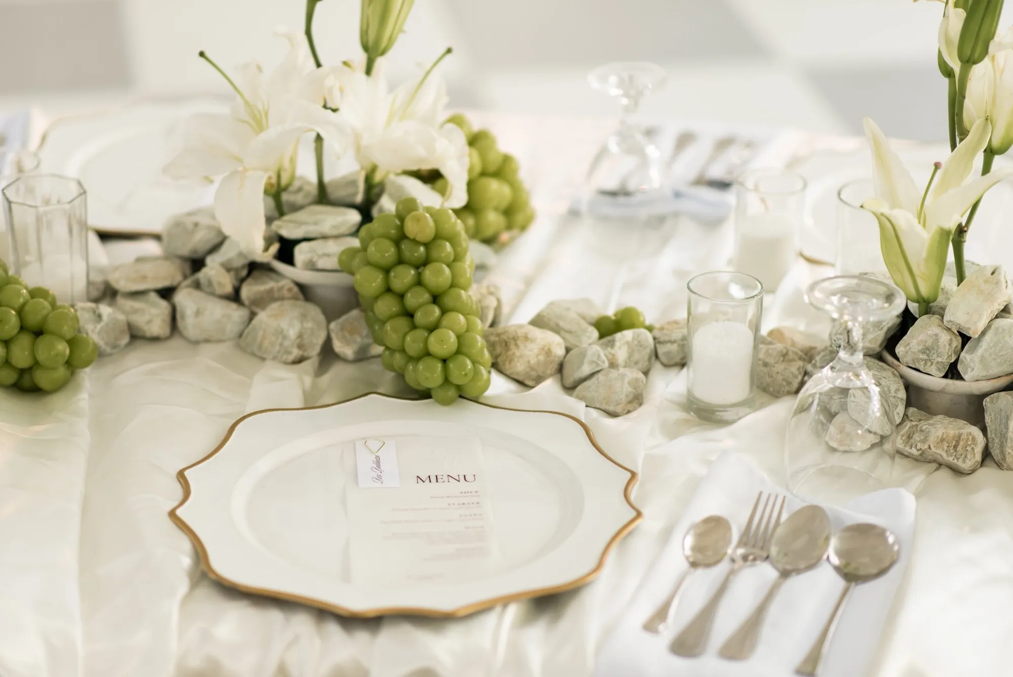 Reception place setting with a gold-rimmed charger, menu card, green grapes, river stones, white lilies, and votive candles on white satin linen.
