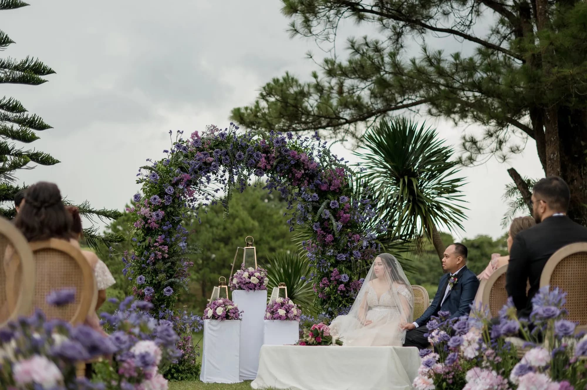 Couple seated before a circular arch of purple and lilac blooms, with lantern-topped white plinths and rose arrangements.
