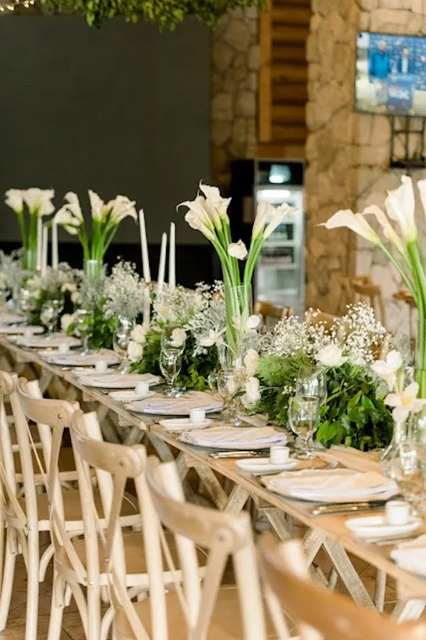 Long wooden farm table with tall white calla lilies, baby's breath, greenery runners, and taper candles, lined with cross-back chairs against a stone wall.