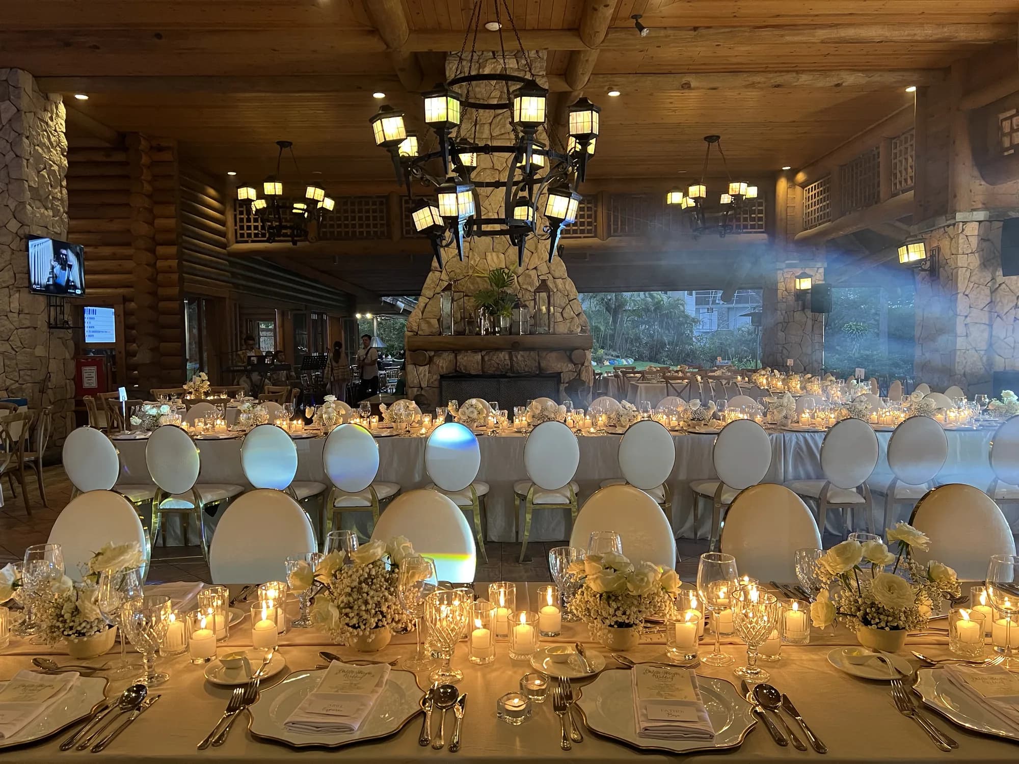 Candlelit reception hall with long white-draped tables, gold-trimmed oval-back chairs, white rose centerpieces, and iron lantern chandeliers framing a stone fireplace.